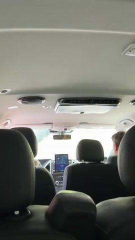 This video provides a rear-view perspective from inside a vehicle, looking forward. The primary subjects are the back of two car seats in the foreground, with a view of the front seats and the road ahead through the windshield. In the middle row, the headrests of two seats are visible, and a person's arm is seen reaching out from between them. The front of the car shows a dashboard with a touchscreen display and a rear-view mirror. Mounted on the ceiling of the vehicle is a climate control unit or entertainment system.

The scene appears to be an outdoor setting, with lush green foliage visible through the windshield, suggesting a rural or natural environment. The lighting indicates it is daytime, possibly late morning or afternoon, with bright conditions filtering into the car. The activity captured is a journey, with the vehicle in motion as suggested by the slight blur of the scenery. The hand gesture could imply communication, adjustment of controls, or simply a relaxed posture during travel. The setting is likely Trnovo Municipality in Bosnia and Herzegovina, as indicated by the location context. No text is clearly visible in the video.
