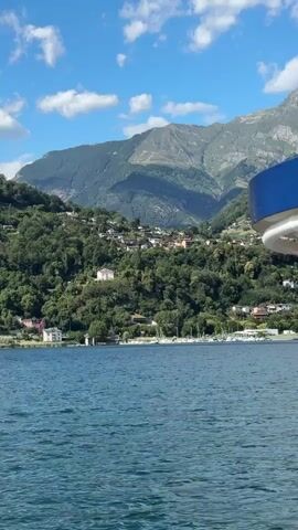 A video pans slowly across a wide shot of a calm, blue lake with a mountainous landscape in the background. The mountains are covered in green trees and scattered with houses, giving the impression of a village built on the hillside. Several sailboats are anchored in the bay. The sky is a bright blue with fluffy white clouds. The right side of the frame is occupied by the edge of a boat, with a blue railing and white structure visible. There are no people or animals visible in the video.
