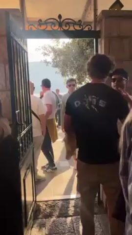 A group of men stand around an open gate on a stone patio on a sunny day. The men wear casual clothes; some wear sunglasses. The gate is metal with ornate, decorative details at the top. Behind them is a sunlit view of trees and water.
