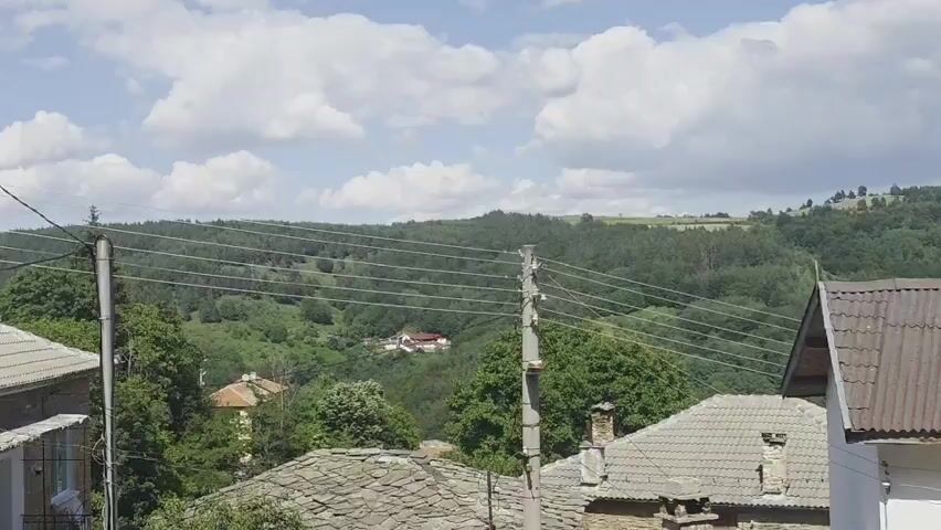 The video pans across a landscape of rolling, tree-covered hills under a cloudy blue sky. In the foreground, several houses with slate and corrugated metal roofs are visible. Power lines stretch across the scene, supported by utility poles. One house in the foreground has a white exterior and a visible chimney. The overall impression is of a rural village nestled in a hilly, forested region.