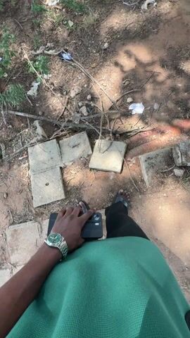 The video is filmed from a first-person perspective, looking down at the ground. The person filming is wearing black sandals, black pants, and a green mesh-textured shirt. A silver watch with a green face is visible on their left wrist, and they are holding a black smartphone with a black case in their left hand.

The ground immediately around the feet is dry, reddish-brown dirt, scattered with several square and rectangular concrete slabs. There are also dried twigs, leaves, and various pieces of litter, including white paper and plastic, on the ground.

The camera slowly pans upward, revealing a wider outdoor scene. Large, leafy trees with thick trunks are prominent, providing dappled shade. In the background, numerous people are visible, mostly young men, standing in small groups or walking across a large, open dirt area. The ground in this wider view also shows scattered litter. To the left, a white building with multiple windows and a dark roof can be seen in the distance, partially obscured by trees. The overall setting appears to be a school campus or a park. The weather is sunny and bright.

The camera then pans back down, returning to the initial view of the person's feet, the concrete slabs, and the debris-strewn ground.