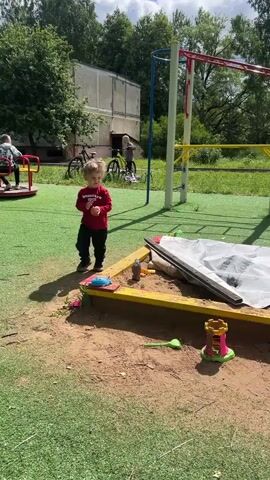 A young boy with blonde hair and a red long-sleeved shirt is standing in a playground. He is holding an orange toy in his right hand and a brown scoop in his left hand. He is wearing black pants and dark shoes. The playground has a green artificial grass surface and a sandpit. In the background, there are trees, a building, and playground equipment including a merry-go-round. The boy is looking towards the right.