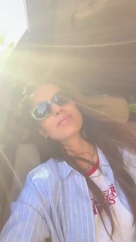 A woman is sitting in a car wearing sunglasses. She has long brown hair and is wearing a blue and white striped shirt over a white T-shirt with red accents. She looks directly at the camera and smiles. The sun shines brightly into the car. She then puckers her lips as if blowing a kiss. The music is loud.
