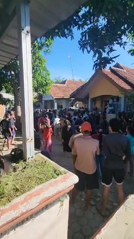 A large crowd of people is gathered in a dirt area between houses in what appears to be a residential neighborhood. The day is sunny, and the sky is clear blue. The houses have tiled roofs. People of various ages and in different clothing are present. Some are standing and observing the scene, while others are moving around. There is a metal ladder leaning against a structure, and a planter box with greenery is visible in the foreground. The overall impression is a community gathering of some kind.