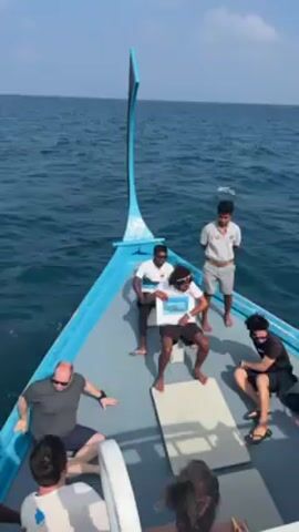 The video shows a group of people riding on a boat in the open ocean. There are about 10 people, including two crew members. The boat is light blue and gray, a traditional Maldivian style boat, sailing on calm waters under a partly cloudy sky. One man in the center of the boat is holding a picture and sits cross-legged on a white cushion. Some people are seated around him, while the crew members are standing.