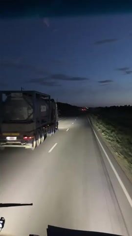 This media file captures a view from inside a vehicle, likely a truck or bus, driving on a highway at dusk. The primary subject is a large truck with a grey cargo container being transported on its trailer, illuminated by the vehicle's headlights. The truck is in front of the viewer, driving on the same highway. The road is paved with white lane markings, and there are fields of vegetation on either side of the road. In the distance, other vehicles with red taillights are visible, suggesting ongoing traffic.

The time of day is clearly dusk, with a deep blue sky dotted with scattered clouds, indicative of twilight. The headlights of the viewer's vehicle are on, casting bright beams on the road ahead. The weather appears clear.

There are no people prominently visible in the frame, though it is implied someone is operating the vehicle from which the footage is taken. The emotional tone is one of solitary travel, perhaps long-distance hauling, with the vastness of the darkening landscape adding to a sense of journey.

No text is visible within the media file itself. The location context provided is Baiqadam, Kazakhstan. This suggests the scene is taking place on a highway in that region, likely during the evening hours. The presence of the cargo truck implies commercial transportation.
