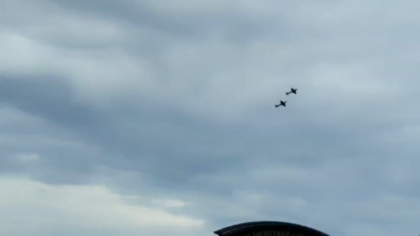 Two airplanes fly in formation across a cloudy sky. A lamppost with a white spherical globe is visible in the foreground on the right side of the frame, and a darker pole with cameras is attached to it.