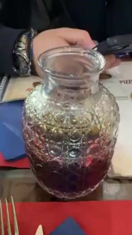 The video shows a table setting in a restaurant, with a focus on beverages. There is a decorated carafe of red liquid, a wine glass filled with red wine, and a bottle of green beer on the table. A blue glass and silverware are also present. The table has a red placemat and a patterned napkin. The video then pans to show the interior of the restaurant. There is a wooden structure with glass panels that displays a QR code for the menu. The restaurant is decorated in a rustic style with wooden furniture and tiled floors. In the initial frame a person’s wrist with a watch and beaded bracelet is visible. Another person is also seen sitting at the table.