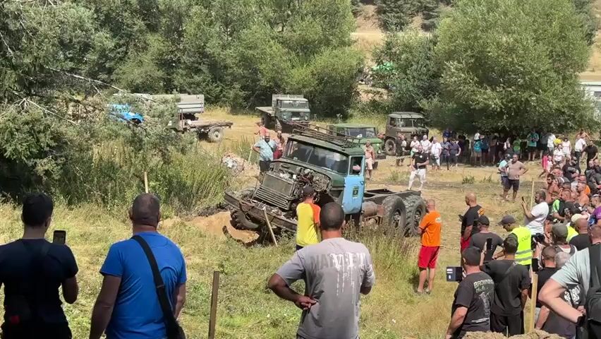 The video shows a large off-road truck that appears to be stuck in a ditch. The truck is green and blue with a large winch on the front. It has six wheels and the front axle is clearly damaged and twisted. The truck is surrounded by a crowd of people, many of whom are watching the truck and taking pictures or videos with their phones. Several other trucks, including military-style vehicles, are parked in the background, some of which appear to be older models. The scene is set in a grassy, wooded area with hills in the distance, suggesting an outdoor event or competition. The lighting indicates it is daytime. The people are dressed in casual clothing.
