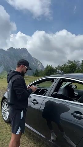 A man wearing a black hoodie, black cap, sunglasses, and black and light blue shorts stands next to the open driver's side door of a black car. He is holding a remote control in his hands and looking down at it. The car is parked on a grassy area. In the background, there are green trees and majestic mountains under a cloudy sky. A drone is visible on the roof of the car.