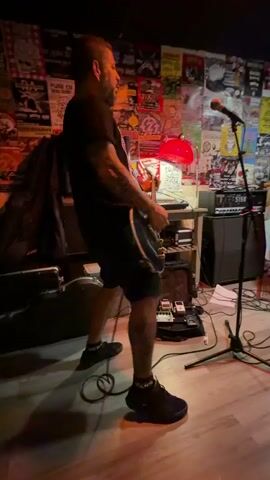 A male guitarist with tattoos on his arms and legs plays a black electric guitar on a wooden stage. He is wearing a black t-shirt and black shorts. The background is a wall covered in posters and flyers. To the left of the guitarist is a guitar case and a dark amplifier. To the right, there is a microphone on a stand, another amplifier, and several guitar pedals on the floor. A red lamp is visible on a shelf behind the guitarist. The scene appears to be a small music venue or practice space. The guitarist is captured in motion, with his body leaning into the guitar.