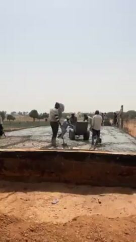 The video shows construction workers pouring concrete onto a large surface reinforced with rebar. The men, dressed in everyday clothes and some with head coverings, use wheelbarrows to transport the wet concrete and then spread it across the surface. The surrounding environment consists of a flat, sandy terrain with sparse vegetation. The sky is bright and clear, indicating a sunny day. The camera is positioned at a low angle, providing a view of the construction site from a slightly elevated position, and remains stationary throughout the video.