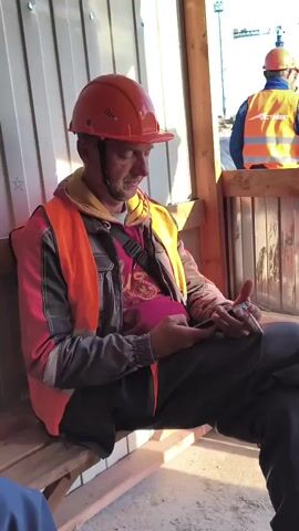 A man in an orange hard hat and orange safety vest is sitting on a wooden bench. He is wearing a pink t-shirt and dark pants. He is holding a cigarette and a phone in his hands. Another man in an orange hard hat and orange safety vest is visible in the background. The environment appears to be a construction site, with corrugated metal walls in the background. The lighting suggests it is daytime.