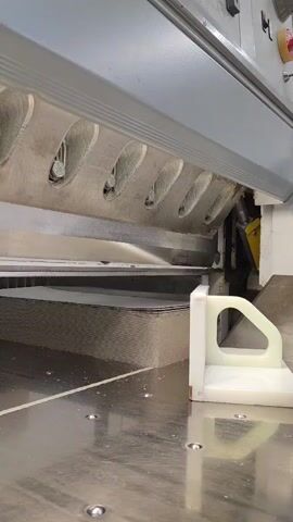 This is a close-up, low-angle shot of a large industrial paper guillotine in operation. The main subject is the cutting mechanism of the guillotine, with its long, sharp blade poised above a thick stack of paper. A white plastic guide or clamp is visible on the right side of the machine, holding the paper in place. The scene is indoors, likely a print shop or a paper processing facility, given the industrial nature of the equipment and the presence of what appears to be paper dust or debris on the metal surface of the machine. The lighting is bright and functional, typical of an industrial setting, and there are no clear indicators of the time of day or weather. No people are clearly visible in the frame, though a hand briefly appears at the very end of the clip, interacting with the white plastic clamp. The emotion conveyed is one of industrial efficiency and power, with the machine poised for a precise cut. There is no visible text in the media file. The location context is Ronnenberg, Germany.