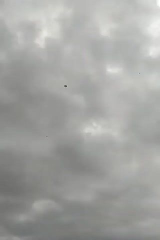 The video shows a dark object descending in the sky under a parachute. The sky is overcast with gray clouds. There are at least two other small, dark objects also visible in the sky. The sound of a siren can be heard in the background.