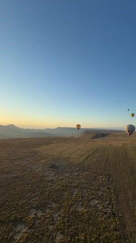 The video captures a scenic hot air balloon flight over a vast, open landscape during sunrise or early morning. The sky is clear and blue, with a warm, soft glow on the horizon. Several hot air balloons are visible in the distance and closer by, gracefully floating above the terrain.

The foreground and midground consist of arid or semi-arid land, featuring fields or plains with a brownish-green hue, possibly with some sparse vegetation. In the background, rolling hills and mountains, some with snow-capped peaks, extend into the distance under the clear sky.

A large white hot air balloon, prominently displaying "TULIP BALLOONS" along with butterfly and tulip designs, is seen closer to the camera, ascending or drifting over the landscape. Other balloons, including a yellow one and a smaller, more distant one, dot the sky. The camera appears to be situated in another hot air balloon, providing an aerial perspective of the breathtaking scenery. The setting suggests a location like Yeşilhisar, Turkey, known for hot air ballooning.