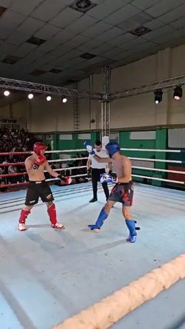 Two male kickboxers are engaged in a match within a boxing ring. The boxer on the left wears a red headguard and shin guards, with black shorts. The boxer on the right wears a blue headguard and shin guards, also with black shorts. A referee, dressed in a white shirt and black pants, stands between them, observing the fight. The ring is illuminated by overhead lights, and spectators are visible in the background, seated in bleachers. The ring itself has white canvas flooring and ropes in red, white, and blue. One boxer throws a punch, and the other attempts a kick. The match appears to be ongoing.