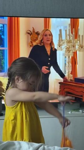 A young girl with dark hair tied in two braids and wearing a yellow sleeveless dress stands in the foreground, looking down. She is holding an object with orange and purple tassels in her hands, manipulating it. Behind her, a large flat-screen television is playing a show. The TV displays an indoor scene with orange curtains, a chandelier, and paintings on the walls. An older woman with blonde hair, wearing a black top and a pearl necklace, is visible on the television screen. As the video progresses, two additional figures, a woman in a black dress and a man in a blue suit, appear on the TV screen. The girl is standing in front of a white piece of furniture, possibly a dresser or TV stand.