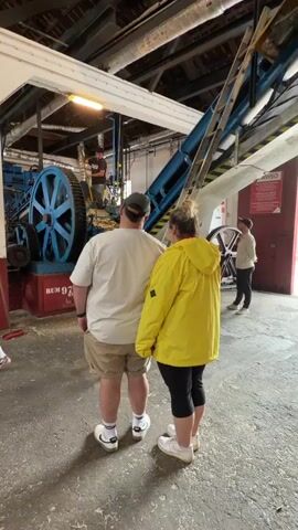 Two people, a man in a white shirt and tan shorts, and a woman in a yellow raincoat and black leggings, stand together in a large, industrial building, looking at a large, blue machine. Another man stands on the machine, and a third man is visible standing near a set of gears on the far right.  There are various pipes and structures overhead. The floor is concrete with wood shavings scattered across it. A red sign with the word "AVISO" is visible on the wall. The base of the machine is painted red with the words "RUM 97" on it. The couple is holding hands.