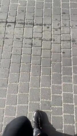 The video shows a person's legs and feet walking on a paved sidewalk. The pavement is made of gray square tiles. The person is wearing black pants, black boots, and striped socks. Sunlight casts a shadow of the person on the ground. The camera angle is looking down at the person's feet. The video transitions to show the person walking on a different section of pavement that appears to be textured with raised dots. The camera remains focused on the person's legs and feet as they walk.