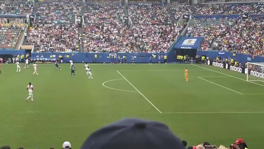 The video shows a soccer match in a large stadium. The field is green grass with white lines marking the boundaries and penalty boxes. Several players in white and blue uniforms are on the field. A referee in an orange shirt is also visible. The stands are filled with a large crowd of spectators. In the foreground, a person wearing a dark blue hat is partially visible, blocking the view of some spectators. The stadium has banners with "FIFA" and "MIAMI" visible. The action appears to be in progress with players moving and interacting on the field.