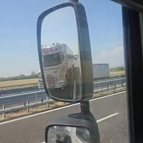 The video captures a view from inside a truck cab, focusing on the side mirror. Reflected in the main mirror is a white semi-truck with red and white detailing and a logo. The truck is on a highway with other vehicles visible in the distance. The smaller, lower mirror reflects the back of the truck the video is filmed from, showing its white trailer and part of the cab. The environment appears to be a sunny day on a highway with dry vegetation along the roadside.