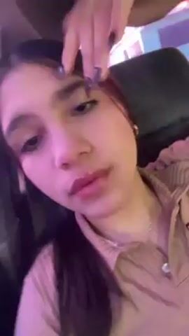 A close-up video shows a young woman with dark hair and fair skin looking directly at the camera. Her face is illuminated with a pinkish light. She is wearing lip gloss. She appears to be resting her face on her hand. The background is dark. The camera angle shifts slightly, revealing a bit of her clothing.