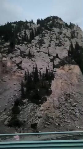 The video is a landscape shot looking up at a rocky mountain slope covered in sparse evergreen trees. The bottom of the frame shows a road with a guardrail. The sky is overcast, and the lighting suggests it is either early morning or late evening. The footage appears to be taken from a moving vehicle.