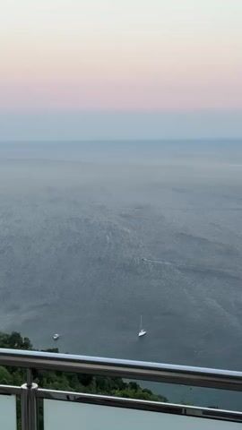 The video shows a view of the ocean from a balcony. The sky is a soft gradient of pink and blue. There are two sailboats visible on the water, one closer and one further away. The water is a deep blue with some rippling on the surface. There is some greenery visible on the left side of the frame, likely from the land. A metal railing is in the foreground, suggesting the vantage point is from a building. The lighting suggests it is either dawn or dusk.