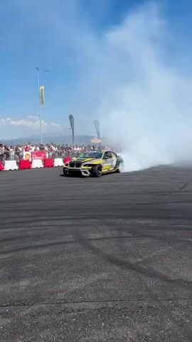 A black and yellow race car with the word "komatsu truck" on its side is shown drifting on an asphalt track, creating a large plume of white smoke. In the background, a yellow car is also drifting, and a crowd of people are watching from behind a barricade. The sky is blue with some clouds, and there are mountains in the distance. The footage is shot from a low angle, capturing the action of the drifting cars.