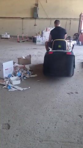 A man is sitting on a green John Deere riding lawnmower with a black bagger attached. He is in a large, empty warehouse-like building with a concrete floor. There are several stacks of wooden pallets to the left of the frame. In the foreground, there is a pile of discarded cardboard boxes and other debris. The man turns his head to look back at the camera. The video pans slightly to the left, revealing more of the warehouse.