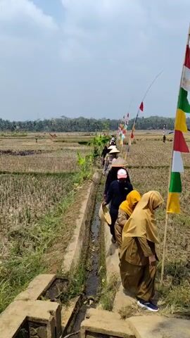 A group of people are walking single file along a narrow path next to a water-filled irrigation ditch. The path is bordered by harvested fields on either side. Many of the individuals are wearing conical hats, typical of those worn by farmers. Some are wearing earth-toned uniforms. Small flags are strung along a pole to the right of the path. The sky is clear and blue.