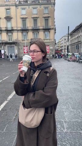 A young woman stands in a European city square, drinking from a Starbucks cup. She is wearing a long brown coat, a grey hoodie, and glasses, with a Calvin Klein crossbody bag. The square is paved with light grey stone, and several scooters and bicycles are parked nearby. There are buildings on all sides of the square, and a few people can be seen walking in the background. The sky is overcast. The woman is facing to the left of the frame, with her arms crossed and the Starbucks cup raised to her lips.