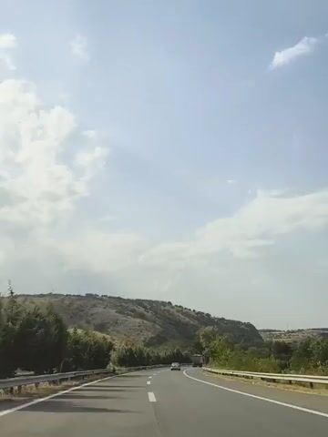 The video shows a view from inside a vehicle driving on a highway. The road curves to the right, and there are vehicles ahead, including cars and a truck. The landscape features rolling hills covered in dry vegetation and scattered trees. The sky is blue with scattered white clouds. The time stamp indicates the video progresses from 00:00 to 00:02.