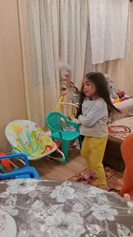A young girl with long dark hair, wearing a light-colored sweater and yellow pants, is actively walking around a room. She is holding a white object in her hand, possibly a sheet of paper or cloth, and is moving it with a flicking motion. The room contains various items of furniture including a table covered with a floral tablecloth, a baby bouncer, a small green plastic chair, and a sofa in the background. The walls are painted in a neutral color and the window is covered with light-colored curtains.