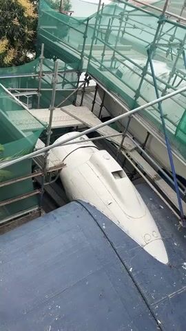 The video shows a close-up, high-angle shot of an aircraft component covered by scaffolding and green safety netting. The component appears to be the nose of an airplane, painted white with some details visible. It rests on a larger, dark blue surface, possibly another part of the aircraft or a platform. The scaffolding is made of metal poles and planks, with the green netting draped over it. The netting has some lettering visible, partially obscured by the structure. The overall scene suggests ongoing maintenance or refurbishment work on the aircraft.