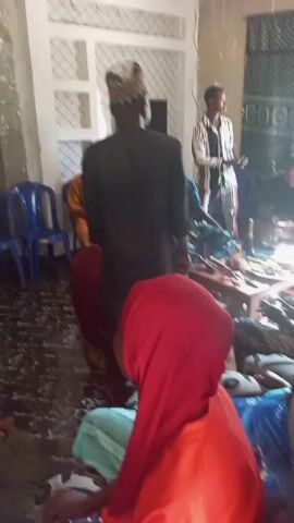 Here is a description of the video:

The video shows a gathering of people inside what appears to be a living room. The room is sparsely furnished with a small wooden table, several blue plastic chairs, and some cushions on the floor. The walls appear to be painted a light color, and the floor has a dark, patterned tile. 

Several people are seated on the floor, mostly women wearing colorful headscarves and dresses. Some individuals are standing, including a man in a black robe and patterned cap, and another man in a white and striped shirt. 

The camera appears to be held by someone moving through the room. The atmosphere seems relaxed and informal, with people gathered in a circle.