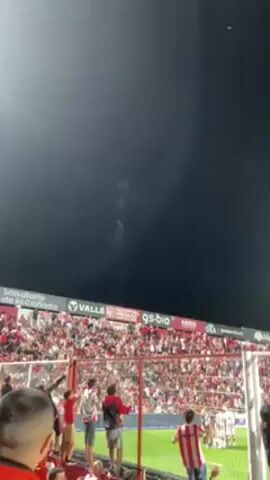 The video shows a stadium filled with people, with fireworks going off in the night sky. The crowd is standing behind a red barrier and appears to be cheering, jumping, and generally celebrating. Some people are wearing red and white striped shirts. In the background, there are advertisements on the stadium walls. The environment is dark, illuminated by the stadium lights and the fireworks.