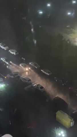 The video depicts a street scene at night during a heavy downpour. The perspective is from a high vantage point, possibly from an apartment building. Cars are parked along the street, and their headlights are visible, reflecting in the rain-slicked pavement. The rain is falling heavily, creating a misty effect. Streetlights illuminate the area, casting a glow on the wet surfaces. Trees line the street, adding a touch of greenery to the urban landscape. A building is visible to the right of the frame. There is a distinct sound of wind and rain audible throughout the video.