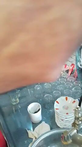 Here is a description of the video:

The video shows a person pouring tea from a white teapot into a small glass cup. The cup is sitting on a saucer with a red and white design. The person is standing behind a table with a variety of items on it, including a bowl of sugar, several glasses, some dirty cups, a bowl, and a damp rag. Through the window behind the table, a blue shirt on a mannequin can be seen. The person is wearing a red shirt.
