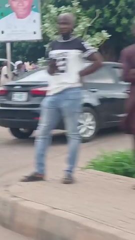 The video captures an outdoor street scene in Hayin Banki, Nigeria, during daylight hours. Several men are gathered along what appears to be a dusty, unpaved roadside or open area. Most of the men are dressed in traditional Nigerian attire, including various styles of kaftans and agbadas, with some wearing hats or caps.

In the foreground to the right, a man in a patterned blue and white garment stands beside another man who is seated on a white plastic chair. Other men are standing around, some appearing to be conversing or observing their surroundings. A red bag is visible on the ground near the center of the frame. On the far left, the front portion of a white van or SUV is partially visible, with more men standing near it.

Overhead, a large green and white billboard dominates the upper half of the shot. The billboard features a prominent image of a man's face, likely a political figure given the common use of such billboards in Nigeria. Visible text on the billboard includes "SAWA" on the left side, and other unreadable text. The overall atmosphere appears casual, with people either waiting, conversing, or simply milling about in the roadside environment. The ground is bare earth, suggesting a less developed area or an unpaved section of the road.