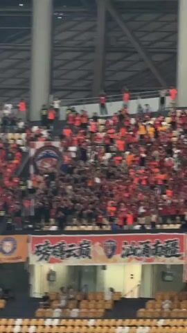 The video shows a large stadium with crowds of people in the stands, many wearing red and waving their arms. Banners are visible in the foreground. The camera then pans down to the field where a soccer team in light blue uniforms is standing. Another player in a yellow uniform appears to be a goalkeeper. An ambulance and other personnel are present near the entrance to the field. The stadium seating is a mix of red and yellow colors.