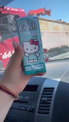 The video shows a hand holding a juice box inside a car. The juice box is a light blue color and has a picture of Hello Kitty on the front. The text on the juice box is in Bulgarian and mentions apple, banana, and carrot. The back of the juice box shows Hello Kitty with two other Sanrio characters. The hand holding the juice box has several bracelets on it. Through the windshield of the car, a red truck is visible in the background. The car's dashboard is also visible, with the radio and air vents.