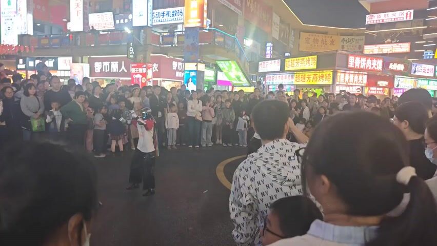 A group of people is gathered in what appears to be a public square or a street in an urban area, likely at night due to the bright lighting. The crowd is looking at two people dressed in costumes, possibly performing or dancing in the center. Many in the audience are holding up their phones to record the event. The surrounding buildings are lit with colorful signs, some with Chinese characters, indicating the location is likely in China. People are mostly standing close together, suggesting it's a relatively crowded event. A person in the foreground is using their phone to capture the scene.