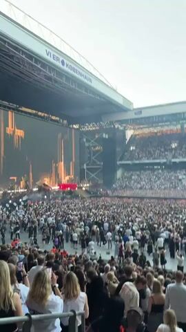 A wide shot captures a concert in a stadium, with a large crowd gathered in front of a stage. The stage is elaborate, featuring multiple levels, intricate set pieces, and large screens displaying visuals. Performers, dressed in red outfits, are visible on the stage, interacting with the audience. The stadium seats are filled with spectators, many of whom are holding up their phones. The overall atmosphere appears energetic and celebratory.