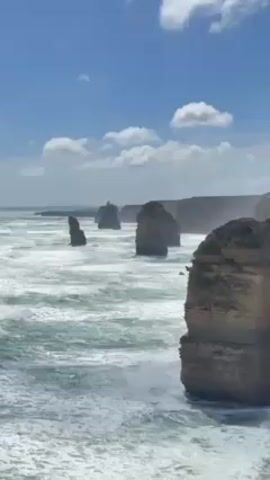 The video displays a view of the Twelve Apostles rock formations in Australia, set against a backdrop of a partially cloudy blue sky. The turquoise ocean is active, with foamy white waves crashing around the bases of the rock stacks. The camera position is static, providing a wide, natural perspective of the coastal scenery.