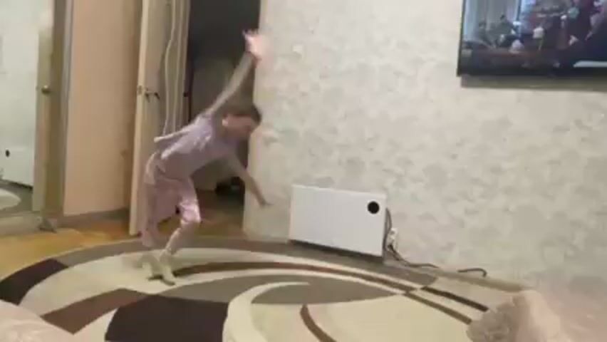 A young girl in a light purple t-shirt and patterned pink leggings is performing gymnastics in a room. She executes a cartwheel and then a series of arm movements and spins, followed by another cartwheel. The room contains a round rug with a curved pattern, a cream-colored couch, a white radiator, a wooden door, and a television mounted on the wall showing a group of people. The girl is the primary focus of the video.