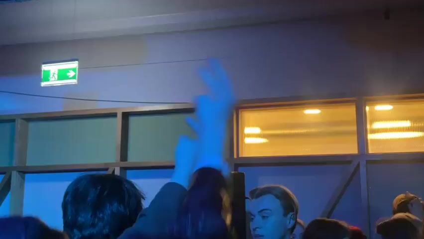 The scene shows the interior of a room under blue lighting, focusing on a crowd with two arms raised in the foreground making a hand gesture with the index and middle fingers extended. A person with short, blonde hair is visible in the crowd. A wooden framed structure with a blue background is behind them, and a window with horizontal lines of light is visible on the right. An illuminated green exit sign hangs from the ceiling.