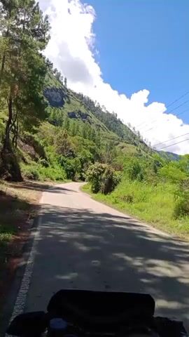 The video shows a first-person view from a motorcycle riding on a paved road that curves to the left. The road is flanked by a steep, green, mountainous terrain covered in trees and vegetation. The sky is blue with scattered white clouds. The sun is shining, casting shadows on the road. There are some utility poles and wires visible on the right side of the road. The lower part of the video shows the handlebars and front of the motorcycle.