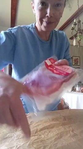 A woman in a light blue sweatshirt is in a room that appears to be a kitchen. She is holding a package of thinly sliced cured meat, possibly ham or prosciutto. She is in the process of opening the package. The meat is pinkish red and appears to be layered. She is holding the package with both hands and her thumbs are inserted into the packaging. The background shows a table covered with a light-colored tablecloth, a chandelier, and some artwork on the wall. There is also crumpled parchment paper on the table in front of her.
